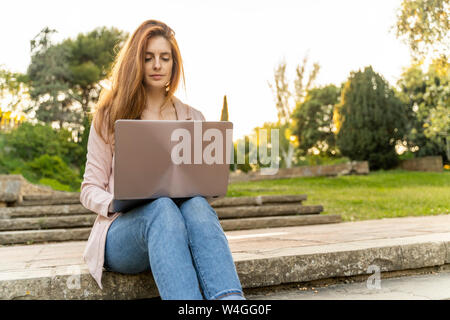 Jeune femme rousse à l'aide d'un ordinateur portable, assis sur les marches dans un parc Banque D'Images