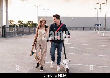 Happy young couple with cell phone, longboard et scooter électrique sur parking Banque D'Images