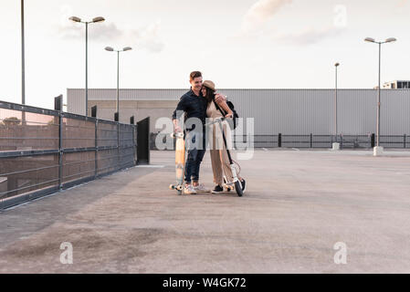 Jeune couple avec longboard et scooter électrique hugging on parking Banque D'Images