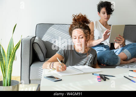 Jeune femme prenant des notes à la maison avec un ami assis sur table à l'aide de tablet Banque D'Images