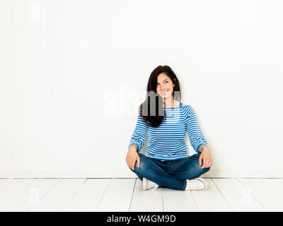 Belle jeune femme avec des cheveux noirs et pull rayé blanc bleu assis sur le sol in front of white background Banque D'Images