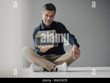 Confiant mature businessman sitting on the floor holding glass house model Banque D'Images