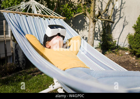Girl lying in hammock en portant des lunettes VR jardin Banque D'Images