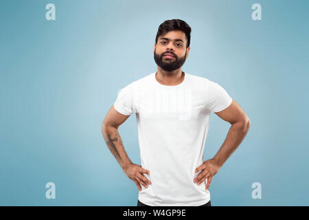 Demi-longueur close up portrait of young man hindou en chemise blanche sur fond bleu. Les émotions humaines, l'expression faciale, ad concept. L'espace négatif. Ce qui pose, debout et souriant, semble calme. Banque D'Images