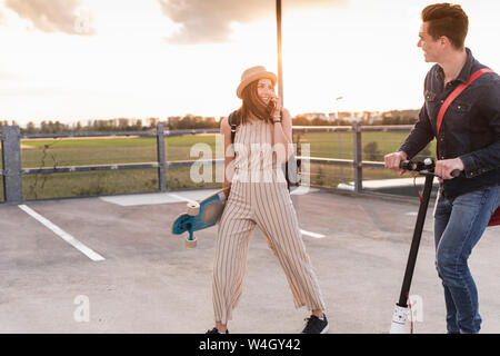 Happy young couple with cell phone, longboard et scooter électrique sur parking Banque D'Images