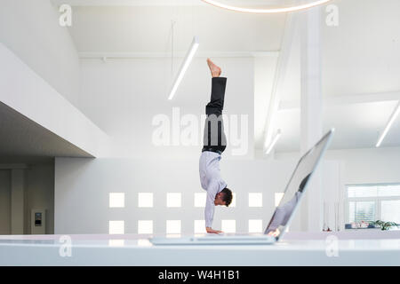 Businessman doing a handstand at laptop in office Banque D'Images