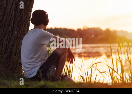 Jeune homme solitaire assis sur le lac par un arbre au coucher du soleil et d'écouter de la musique avec des écouteurs. Un week-end de détente dans la campagne ou le parc. Banque D'Images