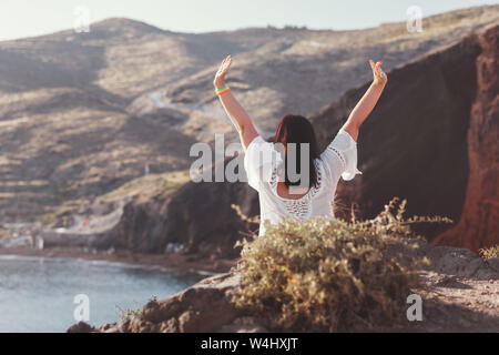 Vue arrière de femme heureuse sur la plage avec les mains, assise sur les rochers de la célèbre plage rouge, avec du sable volcanique et littoral rocheux, Santorin, Akroti Banque D'Images