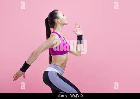 Une jeune brunette jolie jeune fille s'exécute avec le sourire avec des écouteurs sans fil et dans une salle de sport bien haut, sur un fond rose, cardio. Banque D'Images