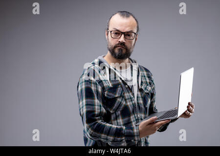 Homme barbu portant des lunettes l'article fixant intensément à l'appareil photo maintenant un ordinateur portable avec une expression perplexe sur isolé gris avec copie s Banque D'Images