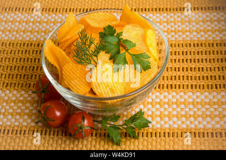 Croustilles de pommes de terre croustillant naturelles dans un bol transparent, décoré avec des feuilles d'aneth et le persil. Près de la cuvette se trouvent trois tomates cerises. Le bol est Banque D'Images
