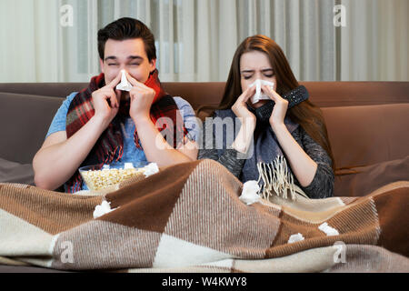 Jeune couple watching movie sur le canapé bel homme et jolie femme portant des foulards soufflant leur nez à la fois à la malade Banque D'Images