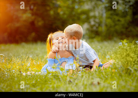 Beaux enfants s'asseoir sur une pelouse verte dans les rayons d'un coucher de soleil. Le garçon des étreintes et des baisers la fille sur la joue. Des enfants heureux Banque D'Images