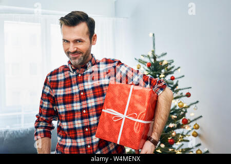 Playful man smiling at camera while holding big cadeau de noël salon décoré dans Banque D'Images