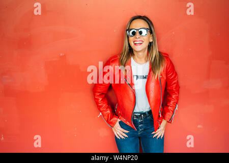 Attractive Woman looking at camera et rire avec des lunettes de soleil et veste de cuir rouge sur fond rouge. Fille de culbuteur en contexte urbain. Banque D'Images