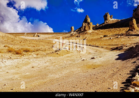 Désert saleté backroad menant à travers la terre sèche de chaud stérile tuf haut spires sur côté droit sous un ciel bleu avec des nuages blancs moelleux. Banque D'Images