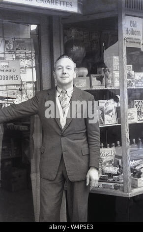 Années 1950, historique, un homme de la presse/propriétaire de jouets dans une veste et cravate debout à l'extérieur de l'entrée de sa boutique, England, UK. Banque D'Images