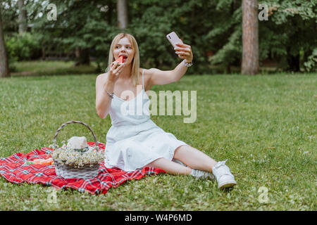 Belle jeune femme assise sur le plaid, eating watermelon, rendant. selfies La nature, pique-nique. Copyspace Banque D'Images