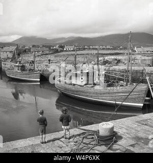 Années 1950, historiques, deux petits enfants debout à un harborl ooking à deux bateaux de pêche en bois, Co Antrim, en Irlande du Nord, Royaume-Uni. Banque D'Images