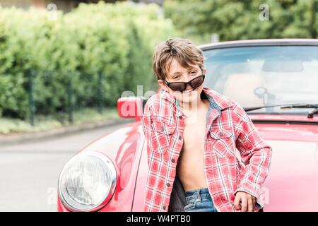 Garçon, 10 ans, avec des lunettes de soleil et chemise à carreaux s'appuie contre une voiture rouge et regarde en souriant à la caméra, Allemagne Banque D'Images