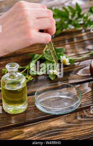 Vue partielle de la femme tenant compte-gouttes près de bouteille avec de l'huile essentielle et bol en verre sur la surface en bois Banque D'Images