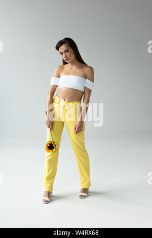 Belle femme en pantalon jaune et blanc haut posant avec le tournesol isolé sur gray Banque D'Images