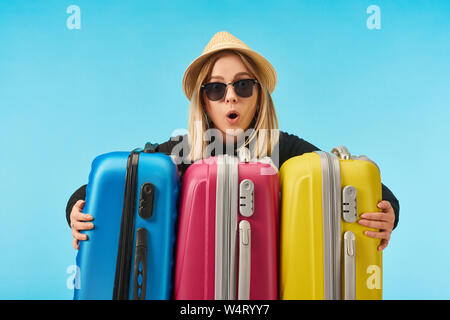 Choqué girl à lunettes de soleil et chapeau de paille près de sacs de voyage multicolore isolé sur blue Banque D'Images