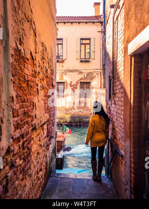 Venise, Italie, 17/11-18. Femme debout sur le bord d'un canal à la fin d'une ruelle. Un vieux classique, bateau, c'est passant. Banque D'Images