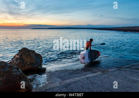 Un homme célibataire paddle boarder entre dans la mer à partir de la cale de halage au coucher du soleil, Westward Ho !, Devon, UK Banque D'Images