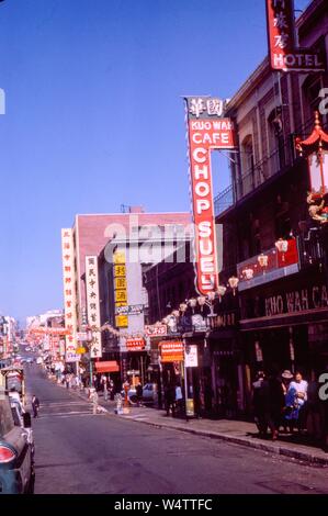 Vue sur la rue de restaurants et boutiques sur Jackson Street dans le quartier de Chinatown de San Francisco, en Californie, y compris Kuo Wah Cafe Chop Suey, Louies de Grant Avenue, et une variété de bannières colorées et enseignes de boutiques, avec les voitures et les piétons aussi visible, image en couleur sur film Kodachrome, 1962. () Banque D'Images