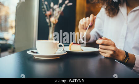 Young woman eating cheesecake et boire du café assis dans une cafétéria près de fenêtre. Close-up de la main de femme à l'aide de fourchette pour obtenir morceau de gâteau. Banque D'Images