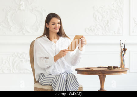 Une belle jeune fille smailing dans un intérieur luxueux est assis sur une chaise, tenant une lettre d'amour de son petit ami et le lit. Sur l'humeur romantique Valent Banque D'Images