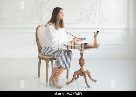 Inspiré d'un artiste fille détient une palette et un pinceau dans ses mains, s'assied sur une chaise dans l'intérieur de l'atelier et donne une image à la table. S Banque D'Images