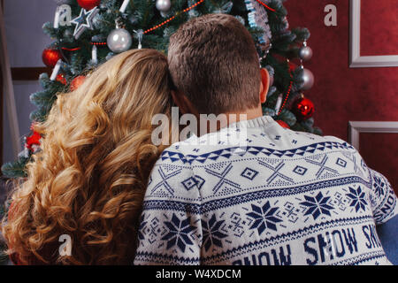 Couple amoureux assis près d'un arbre de Noël, portant des chandails chauds, serrant et à la recherche de l'appareil photo vers l'arbre. Banque D'Images