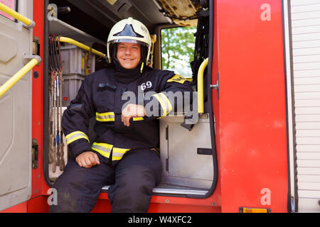 Photo de smiling fireman wearing helmet assis dans un camion de pompiers à la caserne à jour Banque D'Images