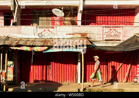 Cuartero, province de Capiz, Philippines : Jeune homme marcher devant les magasins, fermé avec portes pliantes métal rouge, en fin d'après-midi Banque D'Images