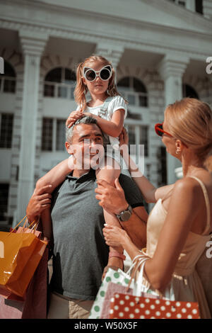 Une sensation de fatigue. De graves little girl wearing big sunglasses sitting om les épaules de son père et de l'écoute de sa mère. Banque D'Images