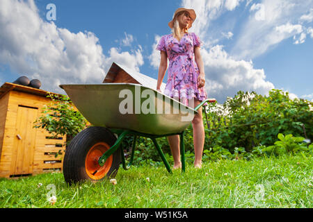 Jeune fille belle blonde en robe et chapeau, s'amuser dans le jardin, tenant dans ses mains un panier vert et à la recherche sur le côté sur la pelouse avec gr Banque D'Images