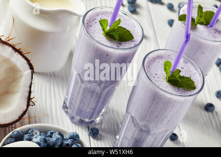 Close-up of blueberry milk-shake mousseux de lait de coco dans trois verres avec paille. Ingrédients sur une table en bois blanc, horizontal Vue de dessus Banque D'Images