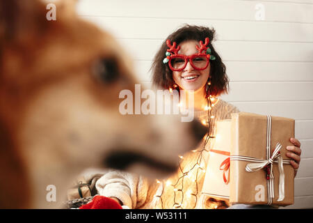 Happy chic girl holding christmas gift box dans les lumières de Noël et souriant pour golden dog cute. Jeune femme en verres de fête avec bois de cerf et Banque D'Images