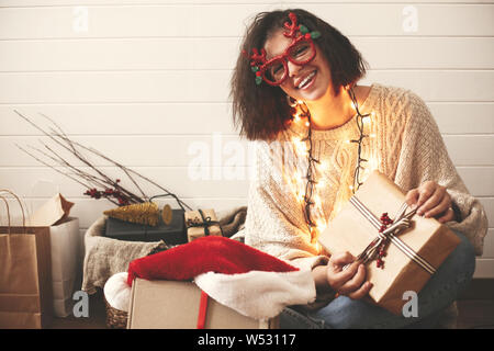 Happy girl élégante dans des verres de fête avec reindeer antlers wrapping christmas gifts and smiling in lumières de Noël. Jeune femme de hipster swea confortable Banque D'Images