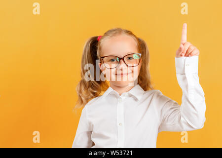 Closeup portrait de petite fille lycéenne sur fond jaune. Un enfant avec des lunettes montre un index vers le haut. Banque D'Images