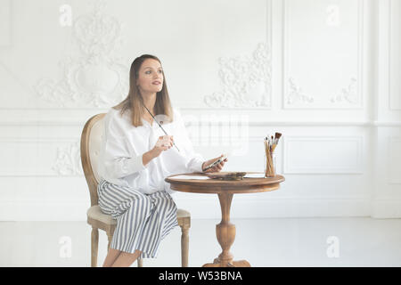 Inspiré d'un artiste fille détient une palette et un pinceau dans ses mains, s'assied sur une chaise dans l'intérieur de l'atelier et donne une image à la table. S Banque D'Images