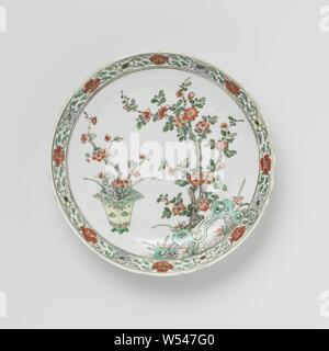 Plat de soucoupe avec des plantes à fleurs dans un pot et à proximité de rochers, plat en porcelaine avec mur de ronde, peinte en bleu sous glaçure et sur l'émail bleu, rouge, vert, jaune, aubergine, noir et or. Sur le plateau sont des plantes à fleurs en pot et près de roches comme le prunus et iris, le mur a un groupe avec motifs floraux stylisés de volutes. Inscrite sur le fond avec une perle dans un double cercle. Quelques jetons dans le bord. Famille verte., anonyme, la Chine, l'c. 1700 - c. 1724, Qing Dynasty, Kangxi (1644-1912) / (1662-1722) Période / Yongzheng-période (1723-1735), Porcelaine, glaçure (matériel), le cobalt (minéral), de l'or Banque D'Images