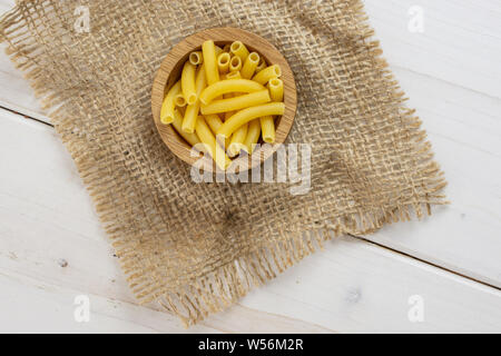 Lot de pâtes crues macaroni morceaux dans un bol en bois avec tissu du jute flatlay sur bois blanc Banque D'Images