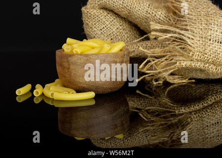 Lot de pâtes crues macaroni dans un bol en bois avec un sac de jute isolé sur verre noir Banque D'Images