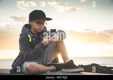 Bel enfant avec des lunettes et black hat skater sitting jouer avec cellphone.Teen reposant, avec téléphone à l'extérieur à l'apprécier le coucher du soleil. Chattin en plein air Banque D'Images