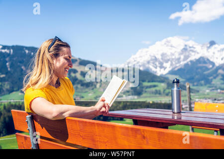 Jeune fille belle et heureuse de lire un livre sur un banc, de montagnes et de verdure à l'arrière-plan. Banque D'Images