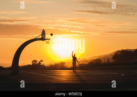 Il joue au basket masculin seul pendant que les couchers de soleil derrière lui à l'extérieur Banque D'Images