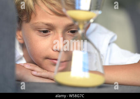 Smiling blonde beau mec allongé avec ses mains sous son visage semble rêveur fasciné en sable lents sablier de verre. rapt regard, concept de de Banque D'Images